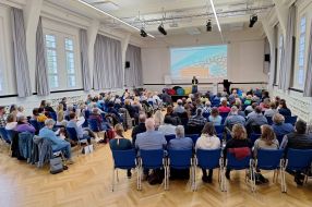 Teilnehmer eines Fachtags lauschen einer Präsentation über wichtige Angebote im Plenum eines modernen Veranstaltungsraums.