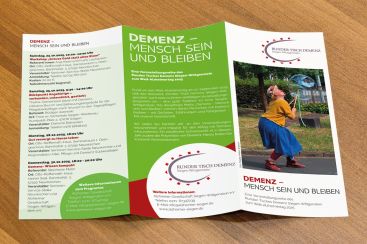 Ein Informationsflyer über die Veranstaltung "Demenz – Mensch sein und bleiben" liegt auf einem Tisch und zeigt bunte Gestaltungselemente.