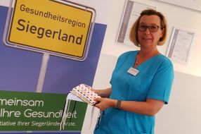 Eine Frau in blauem Kasak hält mehrere Mund-Nasen-Schutzmasken vor einem Schild der Gesundheitsregion Siegerland.
