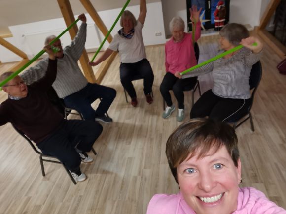 Eine Gruppe Menschen nimmt an einem VHS-Kurs teil und nutzt grüne Fitnessbänder zur Bewegungsgymnastik.