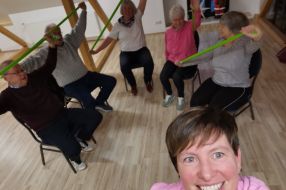 Eine Gruppe Menschen nimmt an einem VHS-Kurs teil und nutzt grüne Fitnessbänder zur Bewegungsgymnastik.