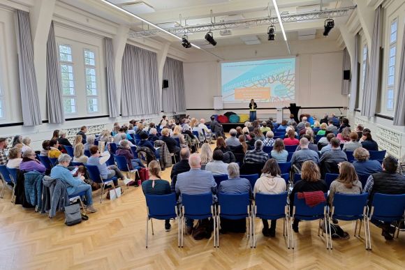 Teilnehmer eines Fachtags lauschen einer Präsentation über wichtige Angebote im Plenum eines modernen Veranstaltungsraums.